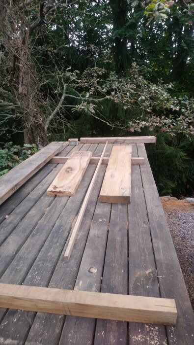 Träplankor och brädor i olika storlekar på ett utomhusbord, omgivet av träd och natur.
