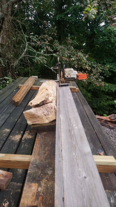 Hyvling och sågning av grov trästock på ett utomhusbord med en motorsåg omgiven av träplankor och grönska i bakgrunden.