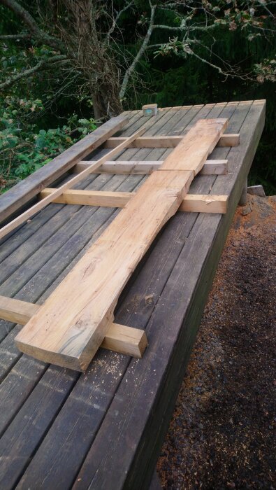 Träplankor och reglar på en arbetsbänk utomhus, omgivna av träd och sågspån.