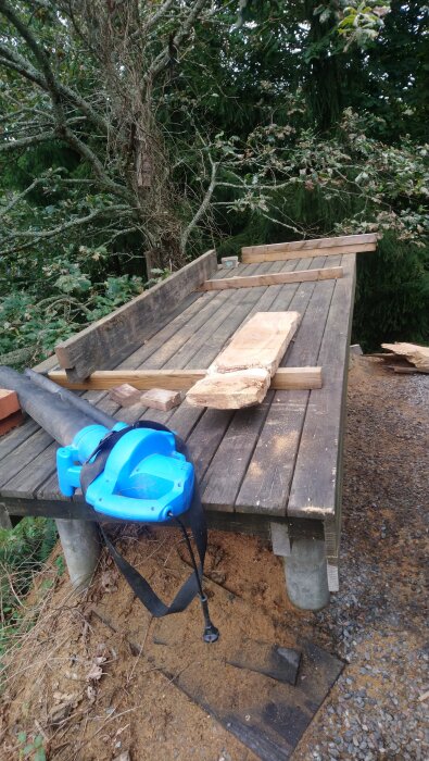 Arbetsbord utomhus med träplankor, hyvlat trä, och en blå elverktygsfläkt, omgiven av träd och natur.