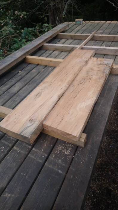 Hyvlade och sågade träplankor ligger på ett trädäck omgiven av grönska, med träspån runtomkring.