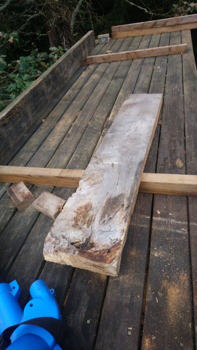 Träplankor ligger på en arbetsbänk utomhus, omgivna av sågspån och blå verktyg i förgrunden.