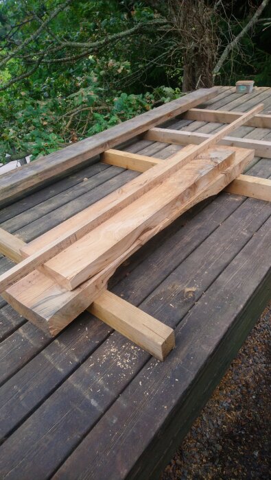 Träbrädor som förbereds för hyvling och sågning på ett utomhusbord, omgivna av trädgrenar och sågspån.