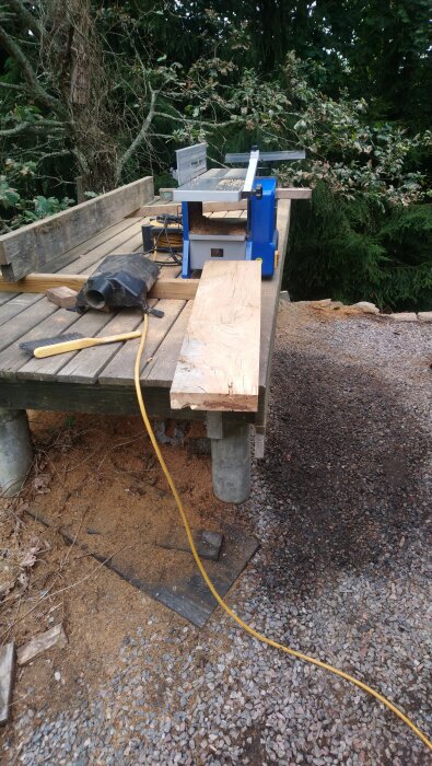 Träbearbetningsområde utomhus med såg, trästycke och borste på ett trädäck, omgiven av grus och skog.