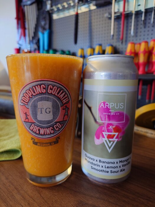 Ett glas med öl från Toppling Goliath bredvid en burk med fruktsmakande smoothie sour ale från Arpus Brewing Co. i en verkstadsmiljö.