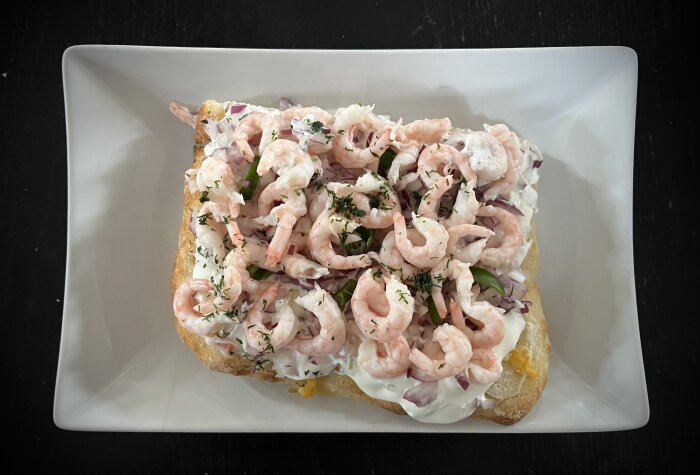 Pinsa toppad med räkor, färskost, mozzarella, rödlök, gräddfil, padrone och dill. Serverad på vit tallrik.