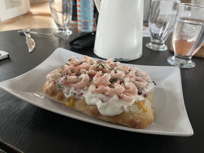 Pinsa toppad med räkor, färskost, gräddfil, rödlök, padrone och dill på en vit tallrik.