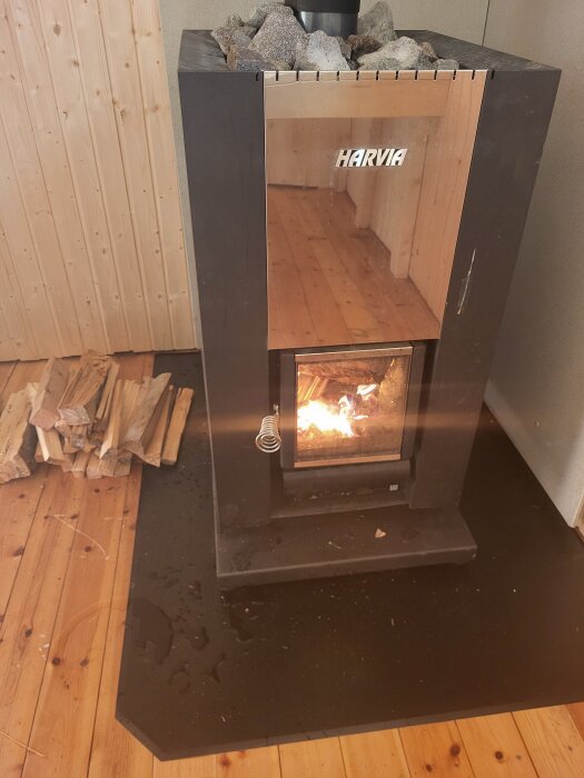 Spis med glasdörr från Harvia, inuti en träklädd bastu med ved bredvid, eld brinner innanför glaset.