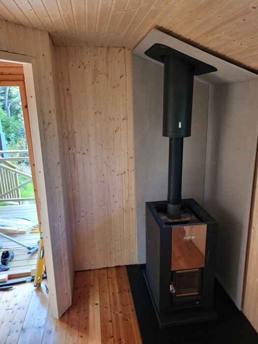 Kamin installerad i trärum med skorsten, trägolv och synlig altan utanför.