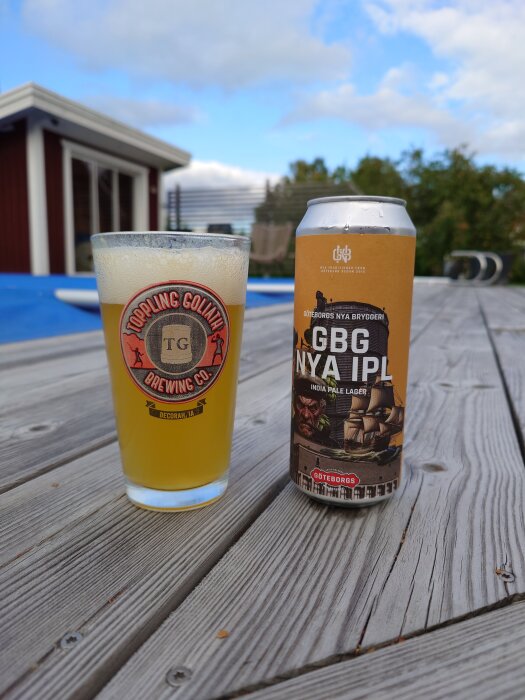 Ett glas och en ölburk på ett träbord utomhus, med en liten byggnad i bakgrunden och en blå himmel med moln.