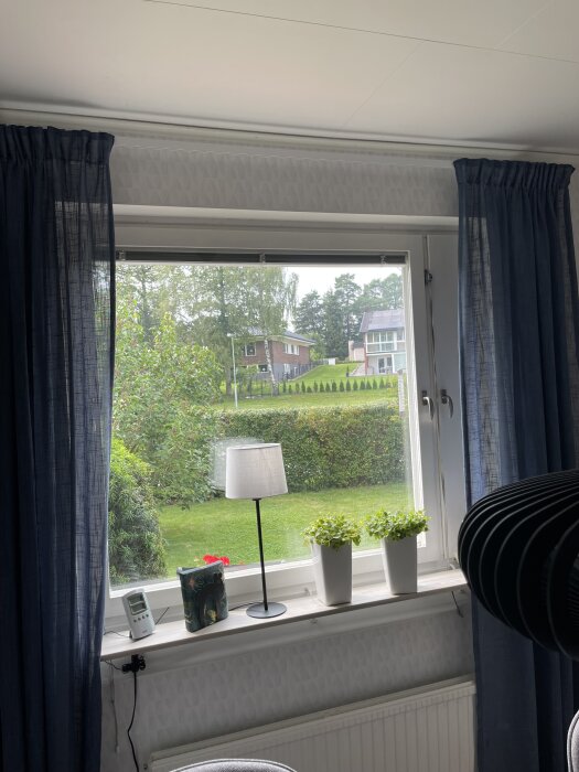 Fönster med tvåglas isolering, vita fönsterbrädor med växter och en lampa, utsikt mot trädgård och hus, blå gardiner på var sida.