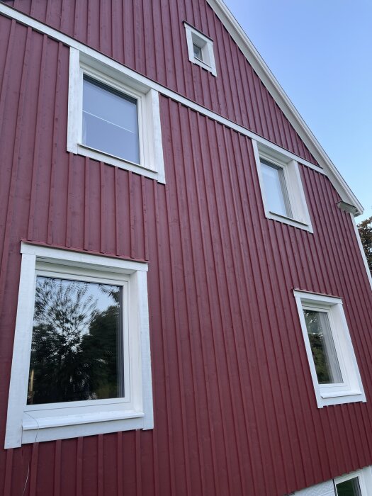 Rött trähus med vita fönsterkarmar av PVC och aluminium som imiterar trä. Fyra fönster syns mot en klarblå himmel.