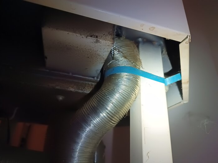 Metallkanal fastsatt med blått band inne i ett utrymme under en Contura kamin, sedd på nära håll, med sotiga ytor synliga.