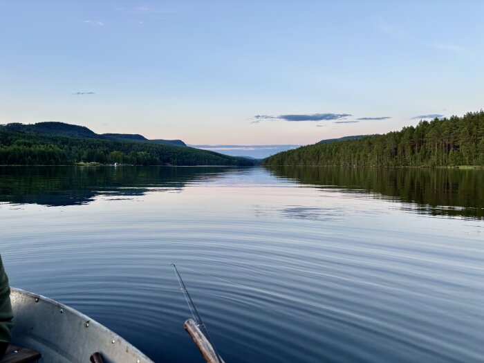 En roddbåt på en stilla sjö omgiven av skog och berg i solnedgångsljus.