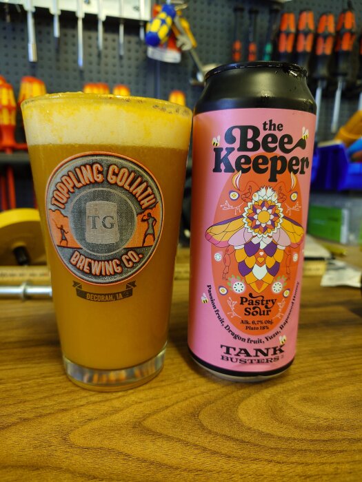 Ölglas med skum från Toppling Goliath och en rosa burk öl med texten "The Bee Keeper" på ett bord, verktyg i bakgrunden.