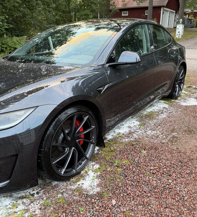 Svart bil på en grusplan efter behandling med skumlans och avfettning, omgiven av grönska och röd byggnad i bakgrunden.