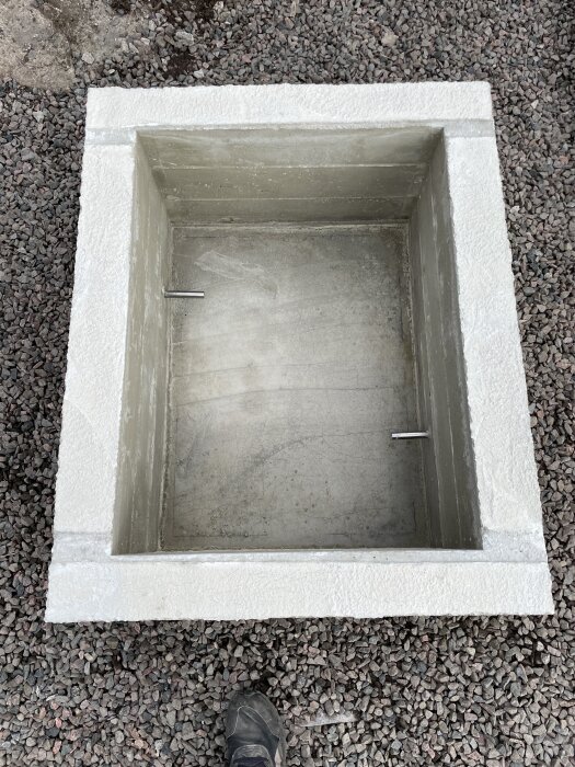 Gjuten fyrkantig betongform med två rostfria stålrör i sidorna, omgiven av grus.