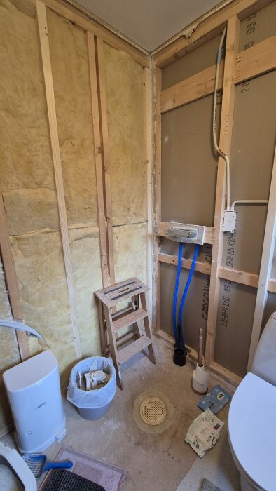 Byggarbetsplats för badrumsrenovering med exponerade väggreglar, isolering, ett avlopp i golvet och diverse verktyg och material.