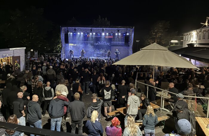 Konsert på kvällstid utomhus med sittande publik vid bänkar och stående folk framför scen där ett rockband uppträder under strålkastarljus.