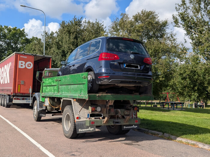 En blå VW Sharan står på ett grönt bärgningsfordon vid sidan av vägen med grönska i bakgrunden.
