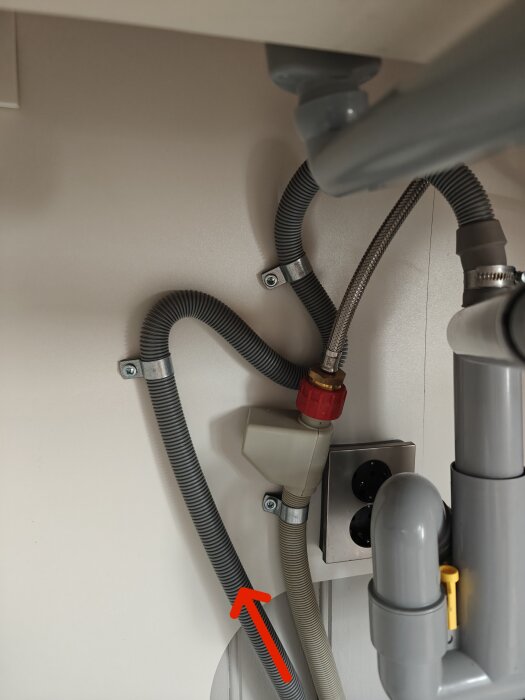 Avloppsslang till diskmaskin markerad med röd pil under diskbänk, visar rör och vattenanslutningar.