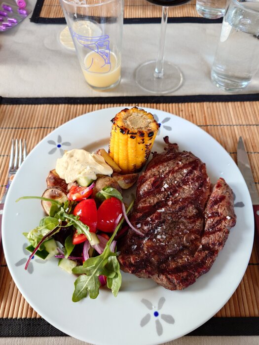 Grillad entrecote och potatis med sallad och majskolv på tallrik, glas juice och vinglas i bakgrunden på dukad bord.