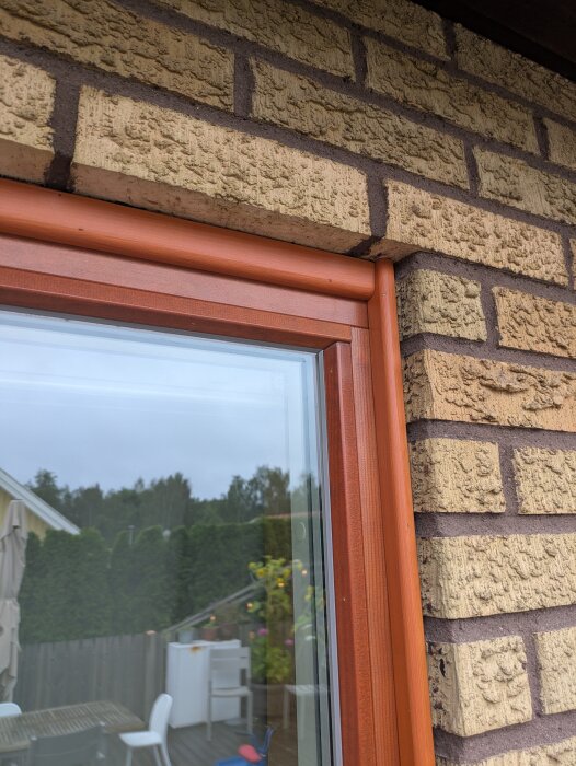 Fönsterram i trä med brunmålad planhyvlad list runt fönster mot tegelvägg.