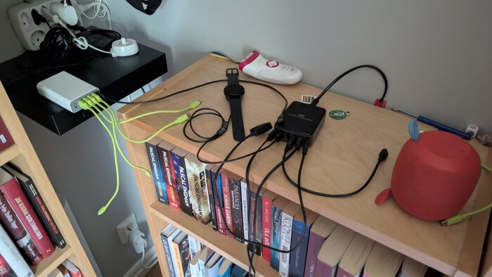 Bokhylla med flera laddningskablar, smartwatch, högtalare och elektronisk apparat. Laddstation med Zigbee-väggbrytare på svart hylla.