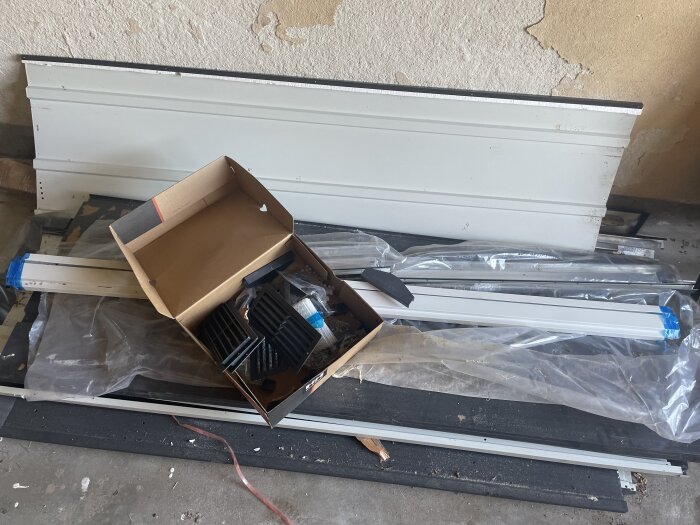 En hög med plåt och byggmaterial på golvet, inklusive en öppen kartong med ventilationskomponenter.