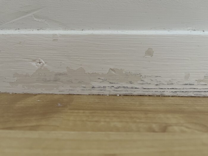 Avflagad färg på en golvlist från 60-talet, där färgen har skrapats bort på vissa ställen och blivit repig, ovanför parkettgolv.