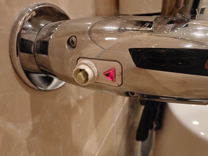 Kromad handdukstork med en avtagna plastbit på vredet och en röd belyst triangelknapp synlig. Möjligen en timer- eller boostfunktion.