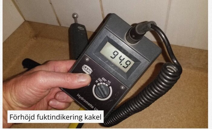 En hand håller en fuktmätare med display som visar 94.9, används för att mäta fukt i kakel.