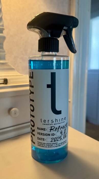 Sprayflaska märkt "tershine prototype refract version 4.0", fylld med blå vätska, står på en ljus träyta inomhus.
