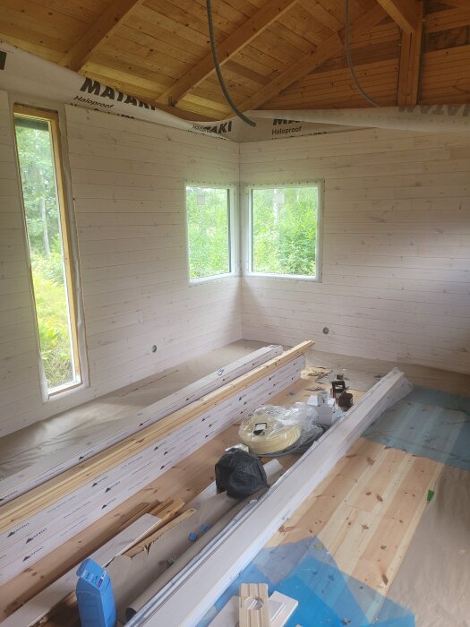 Under renovering i ett rum med synliga träbalkar, två fönster, och staplade byggmaterial på golvet. Träpanel och plast syns också.