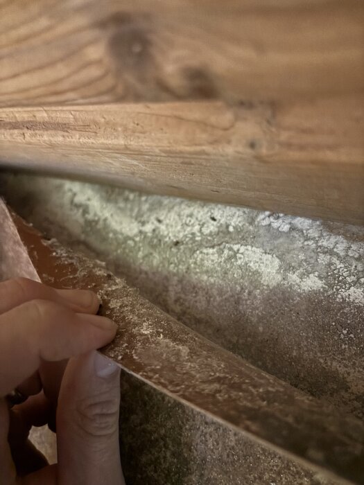 En hand lyfter en gammal plastmatta för att avslöja en vit yta under, potentiellt asbesthaltigt, i en byggmiljö.