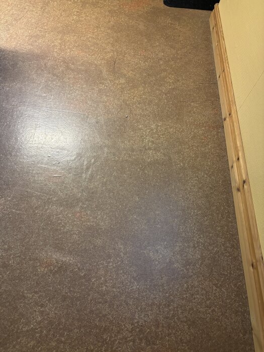 Ljusrödbrun plastmatta med trälist längs väggen, från 70-90-talet, ifrågasatt för asbestinnehåll.