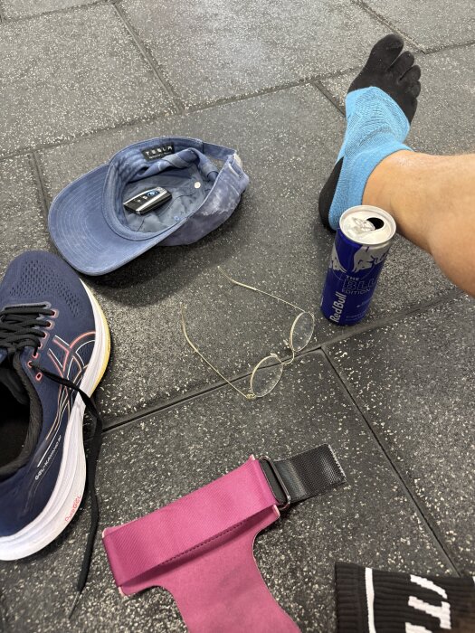 En person med blå tåstrumpor sitter på ett gymgolv med en energidryck, keps med bilnyckel, glasögon, en sko och ett rosa träningsband.