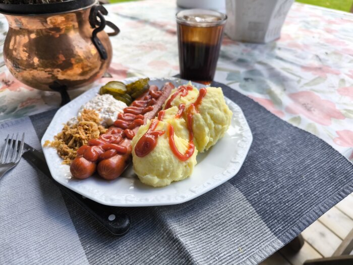 Roslagsgrillare med mos, ketchup, Dallassallad, rostad lök, picklad gurka och ett glas cola på en tallrik utomhus.