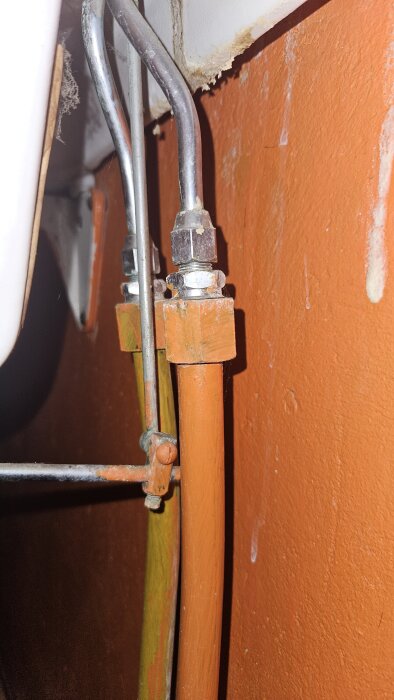 Kopparrör och metallrör med kopplingar under ett tvättställ i ett äldre hus, mot en orange vägg.