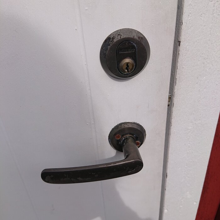 Vit dörr med svart dörrhandtag och ett runt dörrlås ovanför, sett från sidan.