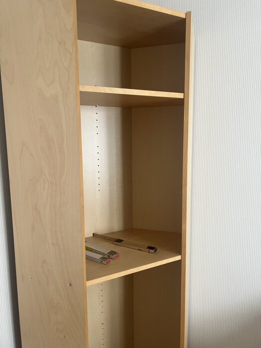 Bokhylla i trä med två hyllplan, en tumstock ligger på det nedre hyllplanet.