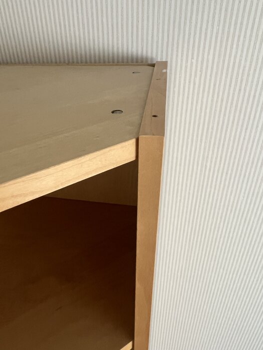 Hörnbokhylla utan dörr, monterad mot randig vägg. Två skruvar syns på hyllans ovansida.