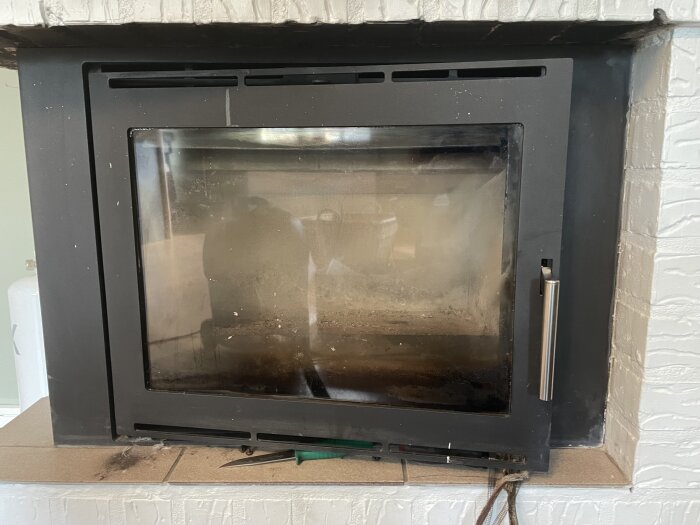 Svart kamin med glaslucka, omgiven av vitt tegel. En skruvmejsel och trasig tätningslist syns på kaminen.