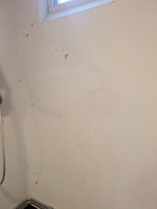 Källarvägg med sprickor och missfärgningar synlig i duschområde, belägen under ett fönster, delvis i behov av ny puts och tätskikt.