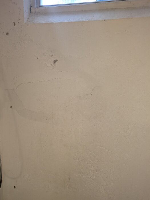 Sprucken och missfärgad källarvägg med vit puts, visar tecken på skador nära fönster i hus från 1973, potentiellt behov av renovering.
