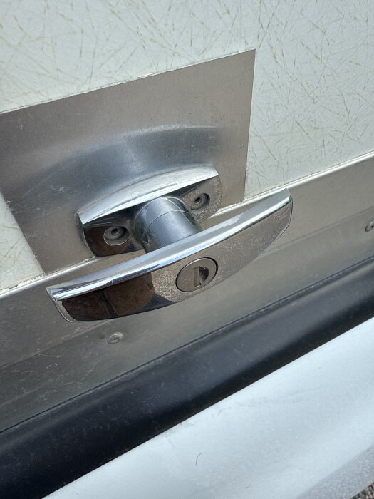 Dörrhandtag i metall monterat på dörr, med synliga skruvhål, potentiellt ämne för utbyte mot ny popnit.