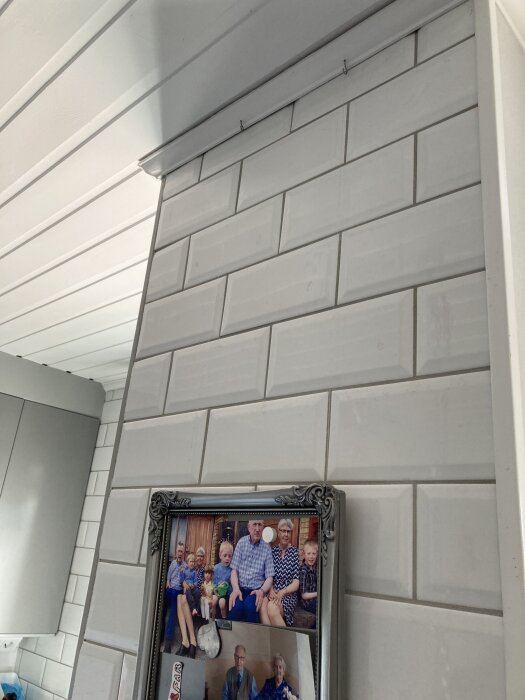 Tavla hänger i fiskelinor från skruvöglor i taklist ovan kaklad vägg, med synliga linor och familjefoto i ram på väggen.