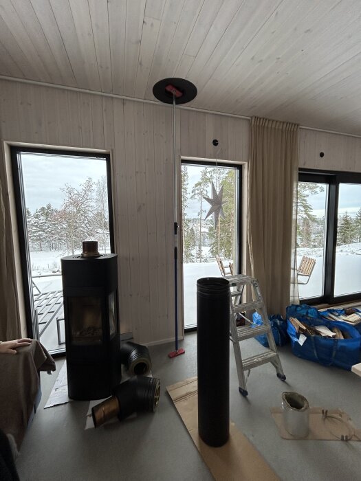 Montering av kamin i ett rum med träväggar, byggmaterial ligger på golvet, två stora fönster visar snöig utsikt.