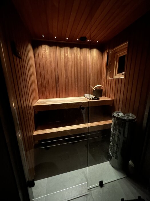 Modern bastu med träpanel, belysning i taket, två bastulavar, en bastuhink och en kamin med stenar.