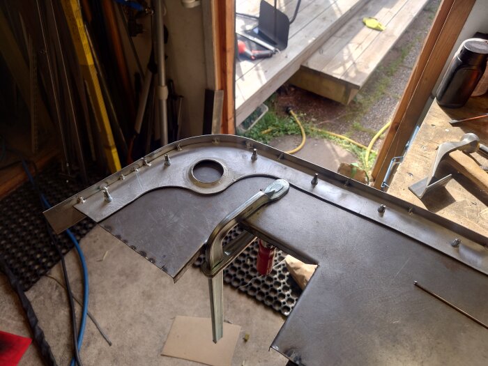 En arbetsstation med en böjd aluminiumplåt klämd fast, omgiven av verktyg och material i ett hobbyrum eller verkstad.